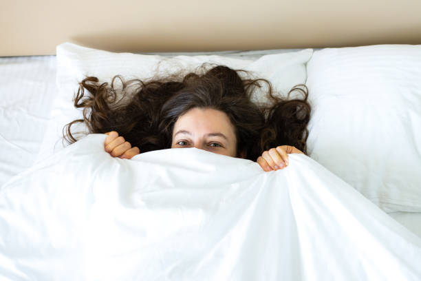 istock Playful young brunette woman hiding face under blanket while lying in cozy bed after waking up, looking at camera, feeling shy peeking from duvet, covering with white sheet. Good morning. Top view 1395202059