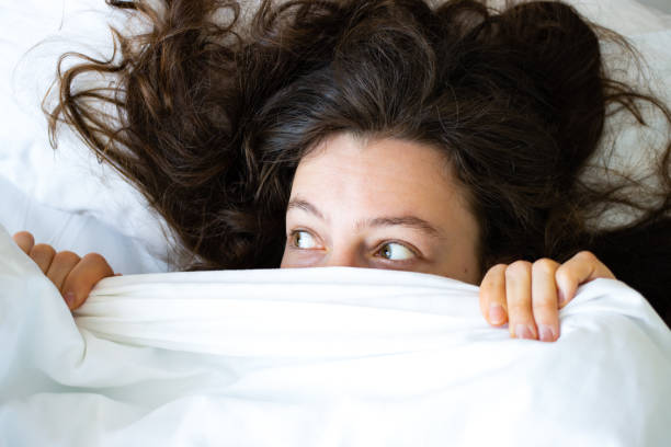 istock Surprised scared young brunette woman hiding face under white blanket, looking in distance, lying in bed. Feeling shy peeking from duvet, covering with sheet. Top view. Scary dream, nightmare concept 1395202062
