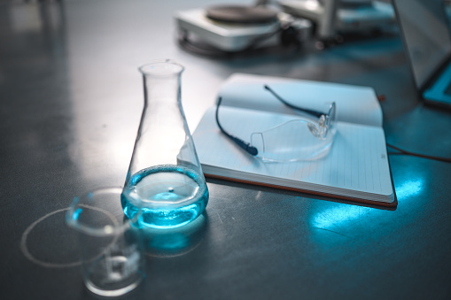 Close up of laboratory glass ware, a glass condenser with blue liquid content and smaller glass jars next to it, notebook on the desk with protective eyewear on top of it. Electric cooker plate in blurred background. Cold light,