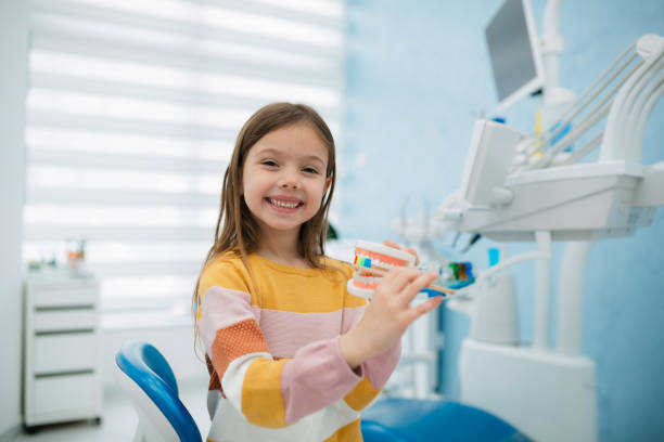 Little girl visiting a dentist Portrait of a happy little girl sitting on a dentist chair and holding a model of human teeth and a tooth brush pretending to brush them, looking at camera dentist' stock pictures, royalty-free photos & images
