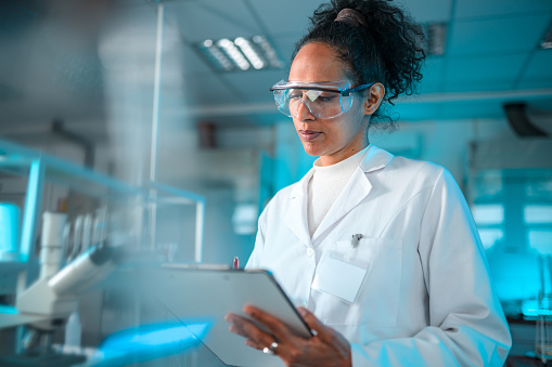 Portrait of a multiracial female chemist in white lab coat with a name tag, wearing protective eyewear, taking notes on a paper pad. Waist up image, a microscope in blurred foreground, various liquid containers and glassware in the background. Looking away. Partly shot through glass.