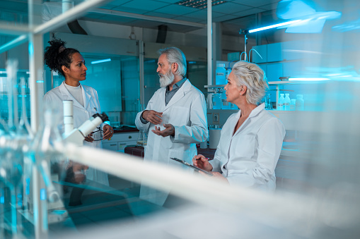 Caucasian female and a male scientists talking to their multiracial female intern. Smiling and discussing something. Older female professional taking notes on a writing pad, side view of his face. Side view of both women's faces. Candid waist up shot through blurred lab shelves.