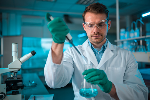 Close up of a male laboratory employee, wearing protective gloves, glasses and a white lab uniform.  Adding liquid substance to a glass condenser with a pipette. Looking away. Blurred background, blueish light.