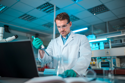 Focused male medical researcher, wearing protective gloves, eyewear and a white coat. Adding liquid substance to a glass condenser with a pipette. Looking away. Low angle view. Blurred foreground and background, blueish light.