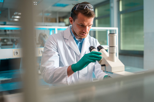 Caucasian mid adult biochemist working with a microscope, dressed in lab coat, wearing latex gloves and protective eyewear on top of his head. Candid waist up shot through lab shelves, looking away.