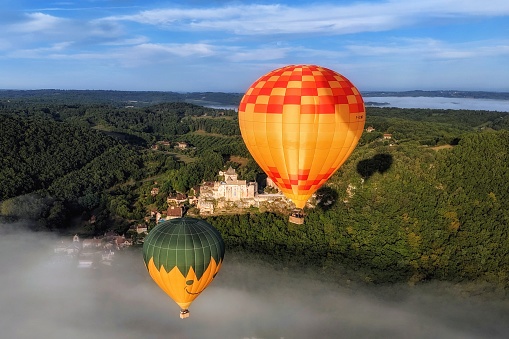 Aerial view of the Chateau de Castelnaud-la-Chapelle, a medieval fortress built on a rocky outcrop, with two hot air balloons flying above the morning mist in the foreground. Périgord, Dordogne Valley, France.