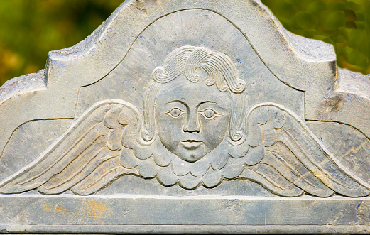 The face and wings of a carved face of a winged angel on an ancient gravestone on Burial Hill in Plymouth, Massachusetts.