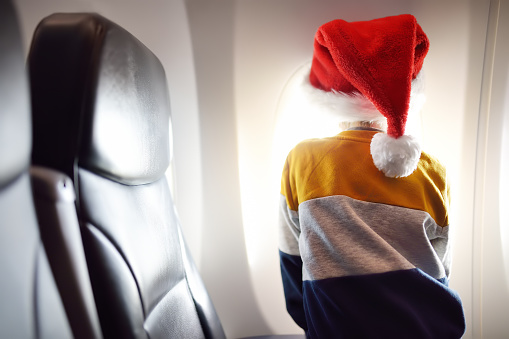 Little child wearing Santa Claus hat is traveling by an airplane. Preschooler boy is looking at aircraft window during flight. Family trip for winter holidays. Happy kid is waiting for merry Christmas