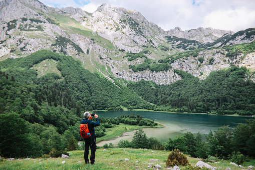 Mid aged man spending a nice day hiking in the nature. He is going across the mountain range of Dinaric Alps in the Southeastern Europe, photographing the mountain lake with his smartphone.
