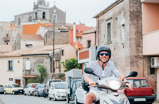 Hombre alegremente sonriente con casco y gafas de sol montando rápidamente el moto scooter en las calles del casco antiguo siciliano con coches estacionados en la Forza d'Agro. Feliz concepto de vacaciones y transporte italiano. photo