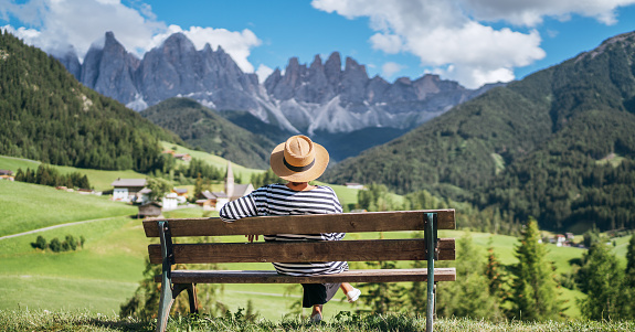 Young fashionable dressed female in straw hat sitting on a bench enjoying Santa Maddalena village view and stunning picturesque Dolomite Alps peaks in Val di Funes valley, South Tyrol, Italy.