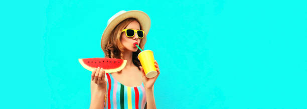 portrait d’été d’une jeune femme élégante buvant du jus de fruits frais avec une tranche de pastèque portant un chapeau de paille sur fond bleu, espace de copie vierge pour le texte publicitaire - watermelon melon fruit juice photos et images de collection