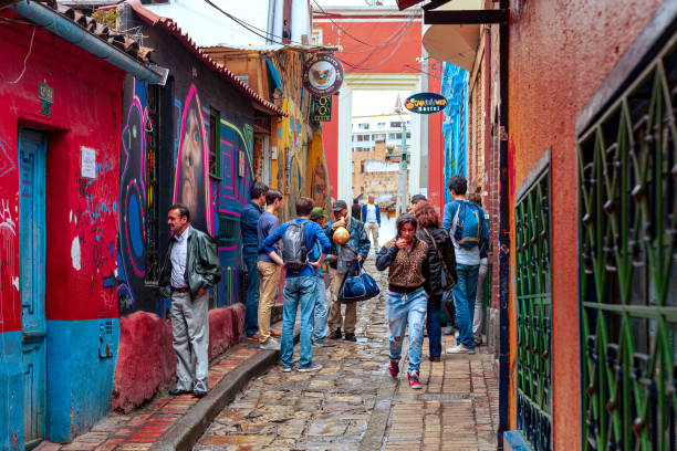 Bogota, Colombia - Tourists And Local Colombians On The Calle del Embudo, In The Historic La Candelaria District Of The Andes Capital City In South America. Bogota, Colombia - July 20, 2016: Both Tourists and local Colombian people walk up the narrow Calle del Embudo one of the most colorful streets in the historic La Candelaria district of Bogotá, the Andean capital city of the South American country of Colombia. The street leads to the Chorro de Quevedo, the plaza where it is believed the Spanish Conquistador, Gonzalo Jiménez de Quesada founded the city in 1538. Many street facing walls in this area are painted with either street art or the legends of the pre-Colombian era, in the vibrant colours of Colombia. A local man tries to sell some souvenirs to a small group of tourists. The altitude at street level is 8,660 feet above mean sea level. Photo shot on an overcast morning; horizontal format. calle del embudo stock pictures, royalty-free photos & images