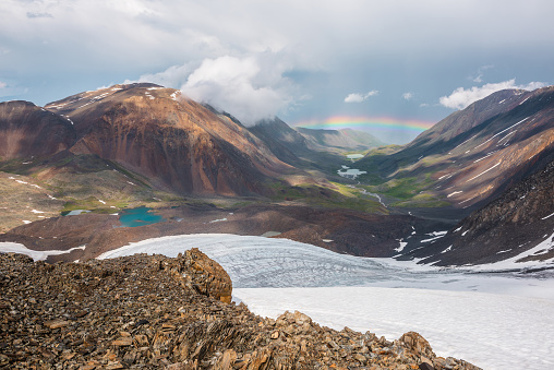 Scenic landscape with vivid rainbow above mountain lake. Alpine scenery with bright rainbow above glacier and glacial lake in mountain valley. Top view to colorful rainbow and low clouds in mountains.