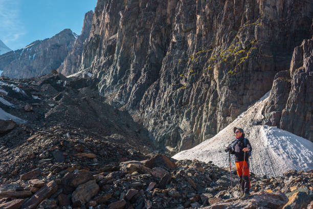Hiker with trekking poles stands on moraine against rocks with glacier in bright sunlight. Man in sunglasses in bright sun near sunlit rocky mountain wall. Tourist in high mountains in sunny day. excursionista con bastones de trekking se encuentra en la morrena contra las rocas con el glaciar a la luz del sol. hombre con gafas de sol bajo un sol brillante cerca de la pared de la montaña rocosa iluminada por el sol. turista en alta montaña en día