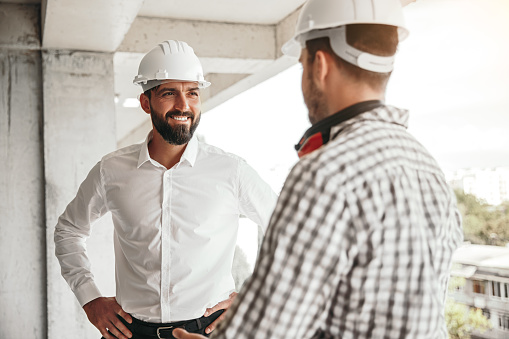 Positive business owner with hands on waist smiling and talking with male builder while checking construction site