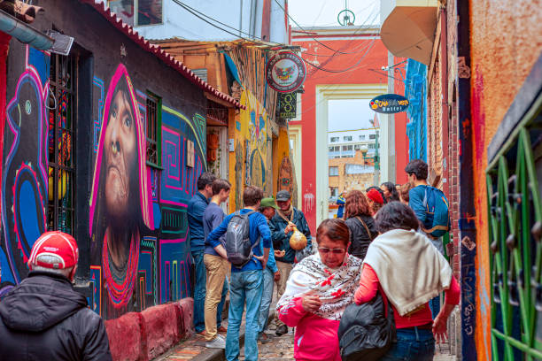 Bogotá, Colombia - Local Colombians And Tourists On The Cobblestoned Calle del Embudo, In The Historic La Candelaria District of The Andes Capital City Bogota, Colombia - July 20, 2016: Tourists and local Colombian people walk up the narrow Calle del Embudo which gets its name from its shape. The English translation of the name would be, "Funnel Street." At this end it is at its narrowest. It is obvious that only two people can walk down the street at this point at the same time. It is one of the most colorful streets in the historic La Candelaria district of Bogotá, the Andean capital city of the South American country of Colombia. Constructed over 450 years ago, when people mainly travelled on horseback, the street leads to the Chorro de Quevedo, the plaza where it is believed the Spanish Conquistador, Gonzalo Jiménez de Quesada founded the city in 1538. Many street facing walls in this area are painted with either street art or the legends of the pre-Colombian era, in the vibrant colours of Colombia. The altitude is 8,660 feet above mean sea level. Photo shot on and overcast morning; horizontal format. calle del embudo stock pictures, royalty-free photos & images