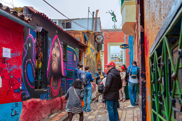 Bogotá, Colombia - Local Colombians And Tourists On The Cobblestoned Calle del Embudo, In The Historic La Candelaria District of The Andes Capital City Bogota, Colombia - July 20, 2016: Tourists and local Colombian people walk up the narrow Calle del Embudo which gets its name from its shape. The English translation of the name would be, "Funnel Street." At this end it is at its narrowest. It is obvious that only two people can walk down the street at this point at the same time. It is one of the most colorful streets in the historic La Candelaria district of Bogotá, the Andean capital city of the South American country of Colombia. Constructed over 450 years ago, when people mainly travelled on horseback, the street leads to the Chorro de Quevedo, the plaza where it is believed the Spanish Conquistador, Gonzalo Jiménez de Quesada founded the city in 1538. Many street facing walls in this area are painted with either street art or the legends of the pre-Colombian era, in the vibrant colours of Colombia. The altitude is 8,660 feet above mean sea level. Photo shot on and overcast morning; horizontal format. calle del embudo stock pictures, royalty-free photos & images
