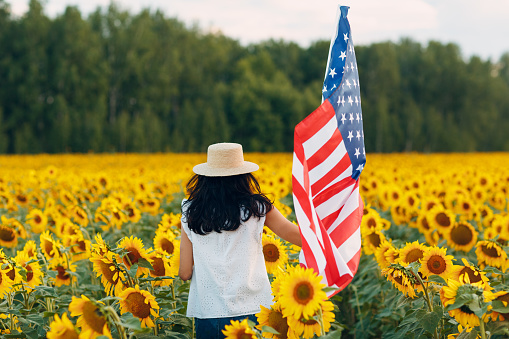 Young woman with American flag in the sunflower field. 4th of July Independence Day USA concept