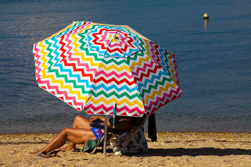 Arousa island, Spain- August 30, 2021: Beach umbrella in front of the sea, woman sitting, sunbathing underneath. Beach, sea water in the background. Arousa Island beach, Ría de Arousa, Rías baixas, Pontevedra province, Galicia, Spain.