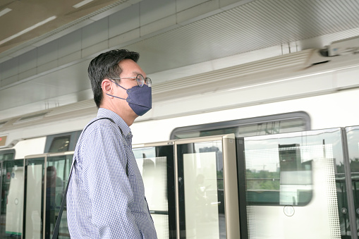 Asian businessman with face mask on standing on subway platform waiting for train. Masked transit concept.