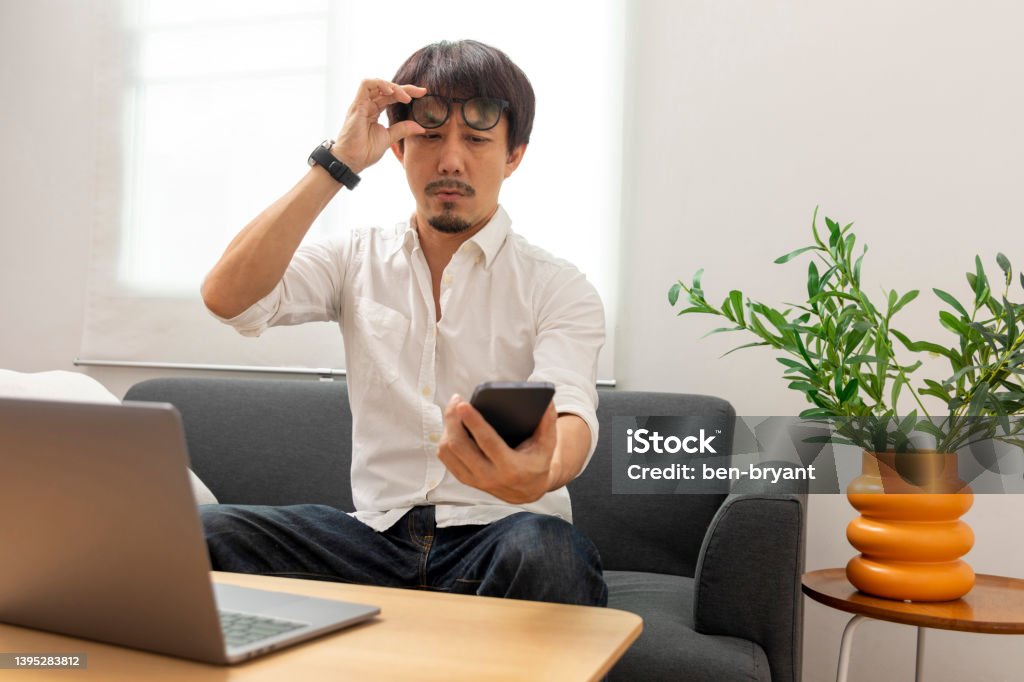 Asian man squint eye to look at the phone due to long eye sighted problems. Middle age Asian man squint eye to look at the phone due to long eye sighted problems, which makes vision difficult. Myopia Stock Photo
