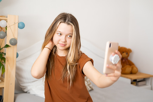 Beautiful Caucasian teenage girl takes a selfie on her smartphone at home, smiles and has fun, admiring himself
