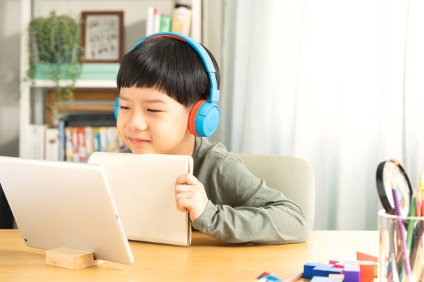 istock Cute elementary Asian student boy showing teacher his notebook in online classroom through tablet. 1395294500