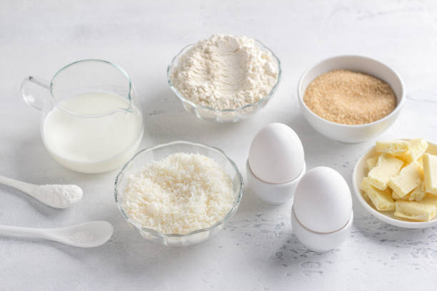 ingredienti per cucinare muffin al cocco al cioccolato bianco: farina, latte o latticello, uova, scaglie di cocco, cioccolato bianco, zucchero di canna, sale e lievito in polvere su un tavolo grigio chiaro - baking flour ingredient animal egg foto e immagini stock