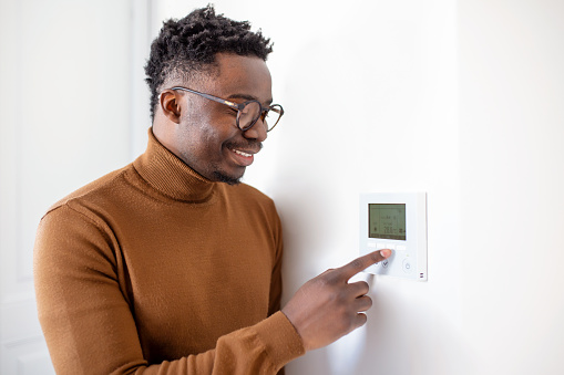 Smiling African American man using modern smart home system, controller on wall, positive young man switching temperature on thermostat or activating security alarm in apartment