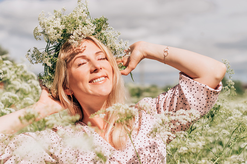 Happy young blonde woman in a pink dress and with a flower wreath on her head in a meadow among flowering grasses.Summer and beauty concept.