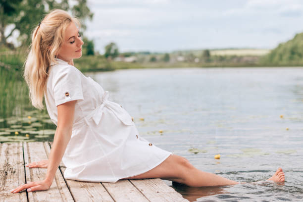 A young blonde pregnant woman in a white dress is sitting on a bridge by the river, with her feet in the water.Summer,pregnancy and prenatal care concept stock photo