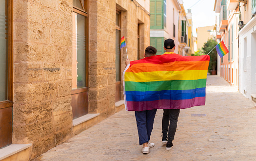 Back view of unrecognizable lgtbq couple in casual clothes and colorful pride flag walking on a city street.