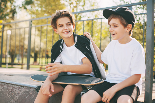 Two cute teenagers sit in a skatepark, relax after skateboarding and chat. Boys enjoy their free time in the skate Park, sitting on the ramp. The concept of youth
