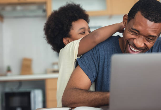 かわいいアフリカ系アメリカ人の男の子がお父さんと抱き合って遊んでいる間、お父さんはラップトップコンピュータを使って自宅で仕事をしていて、現代のキッチンで遠隔作業をしていま� - two parent family indoors home interior domestic kitchen ストックフォトと画像