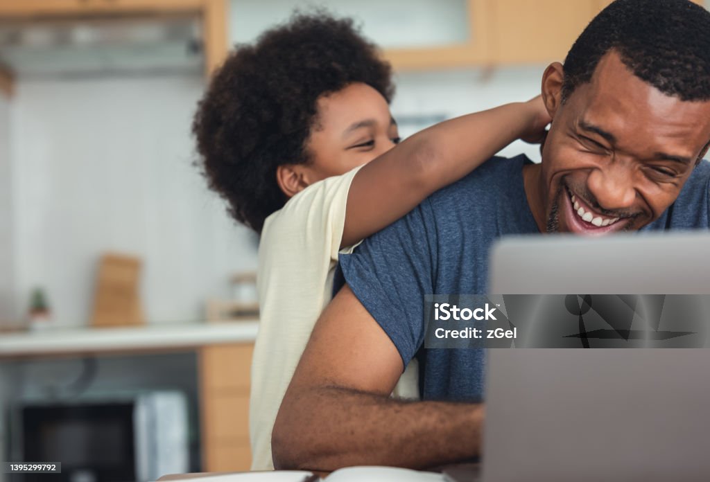 Cute little African American boy hugging and playing with dad while his dad working from home with laptop computer for remoter job in modern kitchen. Concepts of remote work, children, parenthood Father's Day Stock Photo