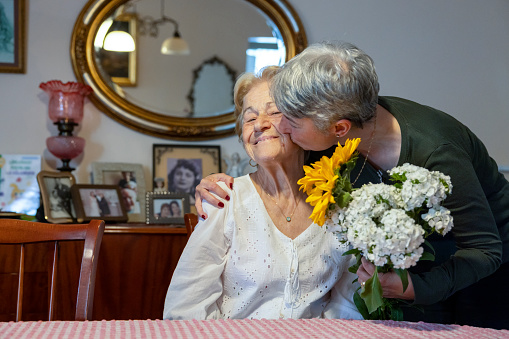 White-haired woman giving flowers and kissing her mother on mother's day at home
