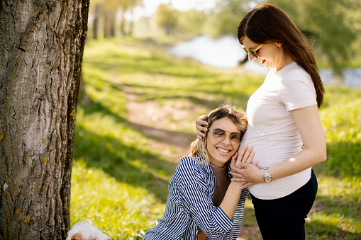 Happy lesbian woman listening her girlfriend's pregnant belly in nature on sunny day, side view