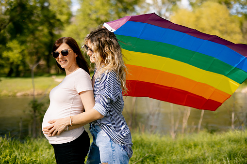 Pregnant lesbian couple with rainbow flag enjoying sunny day in beautiful green nature by the river