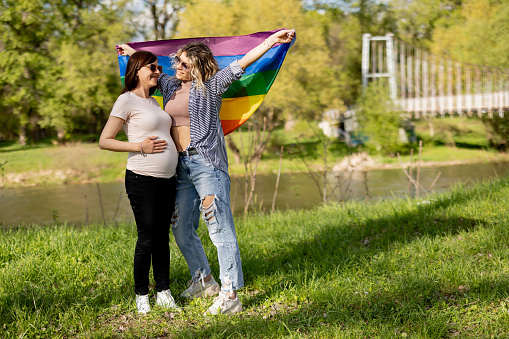 Pregnant lesbian couple with rainbow flag enjoying sunny day by the river