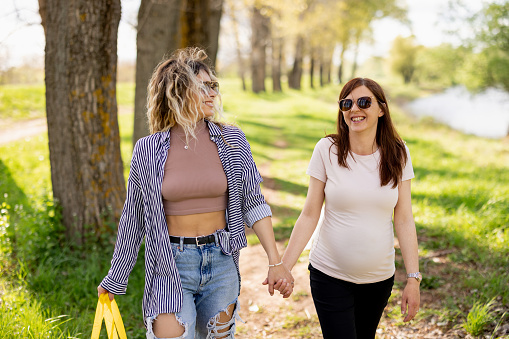 Pregnant lesbian couple with rainbow bag holding hands and walking in beautiful green nature by the river, front view