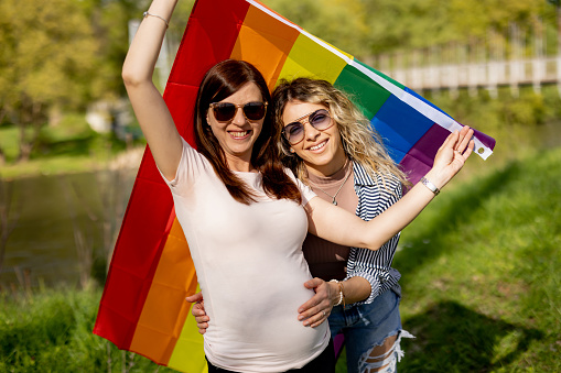 Pregnant lesbian couple with rainbow flag  standing, looking at camera and smiling in green nature on sunny day
