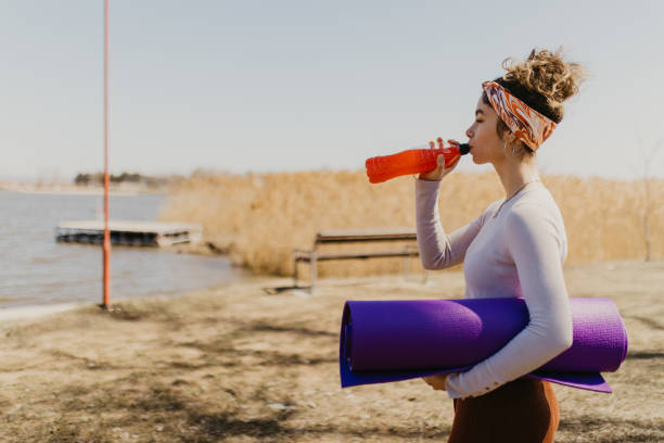 Side view of a young woman with a yoga mat, drinking juice outdoors stock photo
