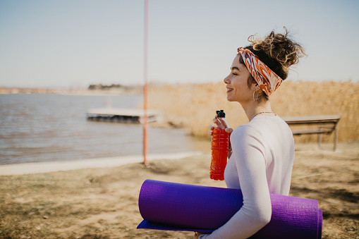 Side view of a young Caucasian woman with a bottle of juice and a yoga mat in nature.