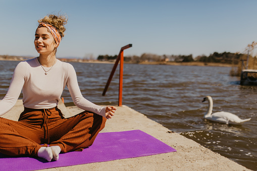 Young Caucasian woman enjoying doing yoga on a sunny day, outdoor by the lake.