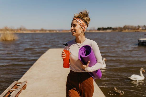 Front view of a smiling young woman with a bottle of juice and a yoga mat in front of the lake stock photo