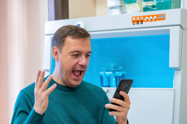Excited millennial scientist showing emotions of joy about victory in online lottery in room with laboratory box in UV light for sterilization in background. Amazed successful male worker of scientific laboratory in casual wear win money celebration success and sit by laboratory shelf inside office, sport betting concept. científico millennial emocionado que muestra emociones de alegría por la victoria en la lotería en línea en la habitación con caja de laboratorio en luz uv para esterilización en el fondo. asombrado exitoso trabajador masculino de laboratorio cientí