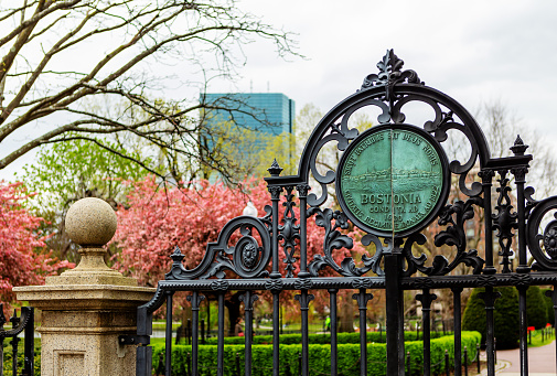 Boston, Massachusetts, USA - April 27, 2022: Close-up of Boston Public Garden iron East gate, Charles Street entrance. Early Spring view of Garden with Back Bay Hancock Tower skyscraper in the background.