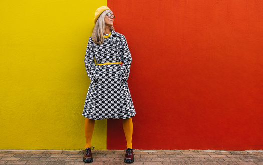 Mature woman looking away thoughtfully while standing against a colourful background. Pensive elderly woman reflecting on memories of the past. Senior woman wearing colourful clothing outdoors.