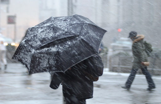 This is a photograph of a pedestrian in a snow storm with an aumbrella with a person in the background without an umbrella.